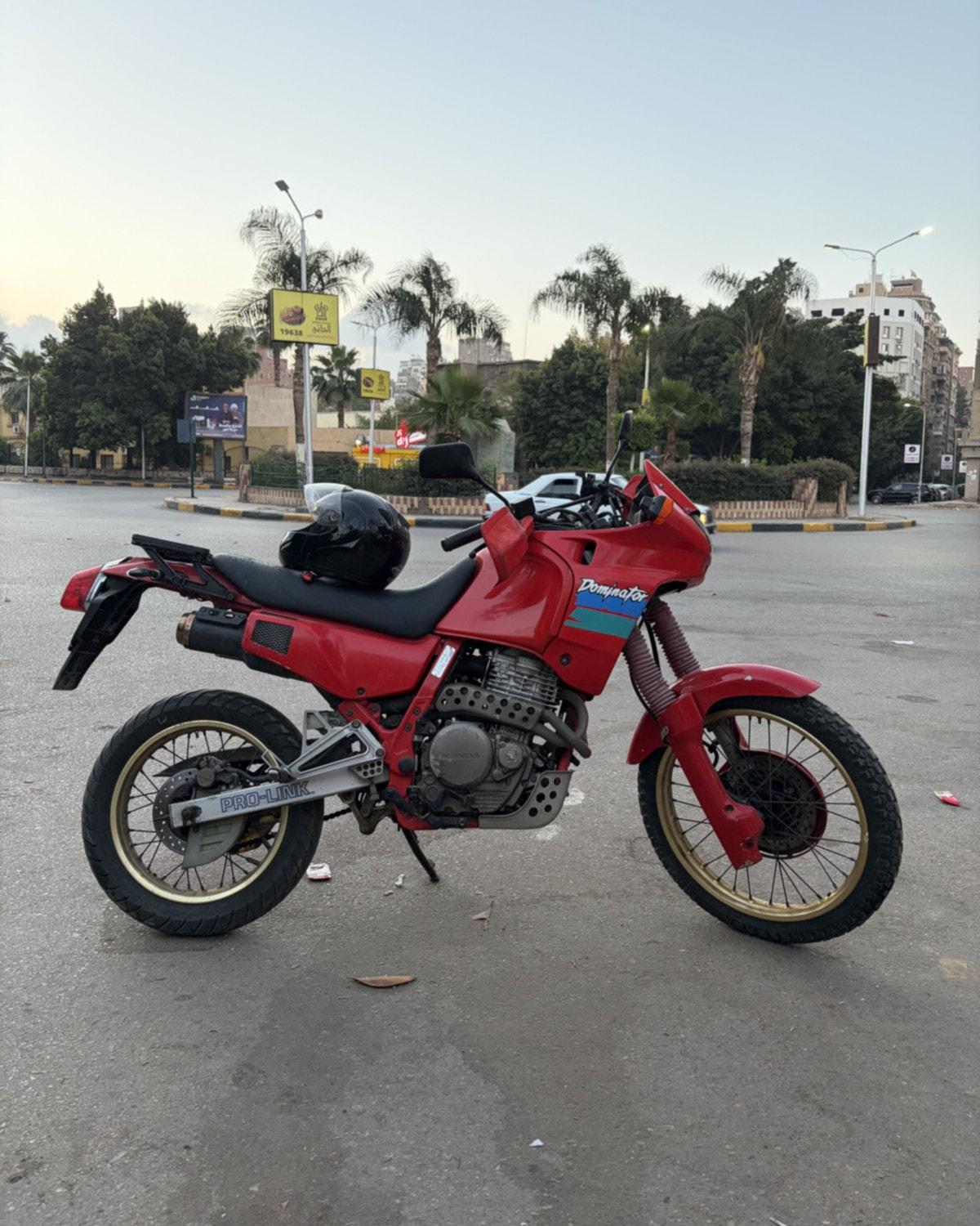 A red 1989 Honda Dominator motorcycle with a black helmet on the seat, parked on an empty street
